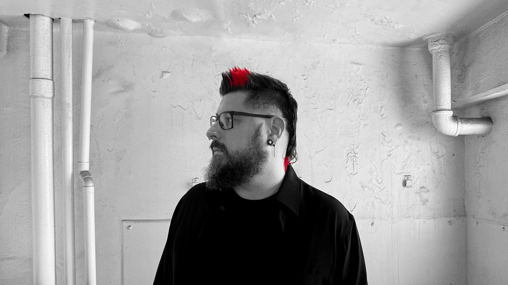 Photo of Yann Novak in a dirty white room with pipes on either side of him and white pain sagging off the walls. He is centered in profile looking to his left and wearing black cloths. The photo is in black and white accept for red accents in his hair.