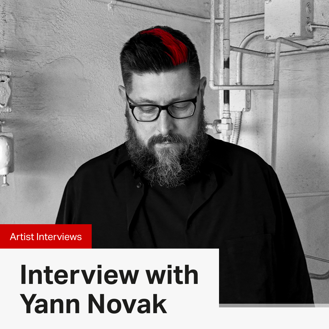 Photo of Yann Novak in black and white with his patches of bright read hair in color. He is wearing glasses, looking down, and wearing a black shirt. The background is a white corner of a room with dirty pipes in the upper right corner. There is text overtop that reads: Artist Interview, Interview with Yann Novak.