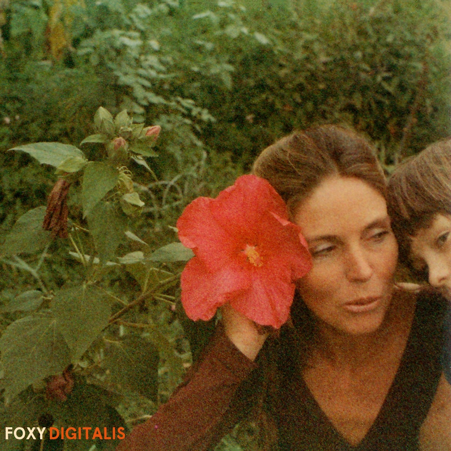 A photo of Ingrid Buslee with her hair pulled back and wearing a long sleeve v-neck shirt with plants all around her and holding a red flower the size of her face just to the left of her face. A very young Yann Novak is peaking his head into frame on the right and Ingrid is looking at him. In the bottom left it read 'Foxy Digitalis.'